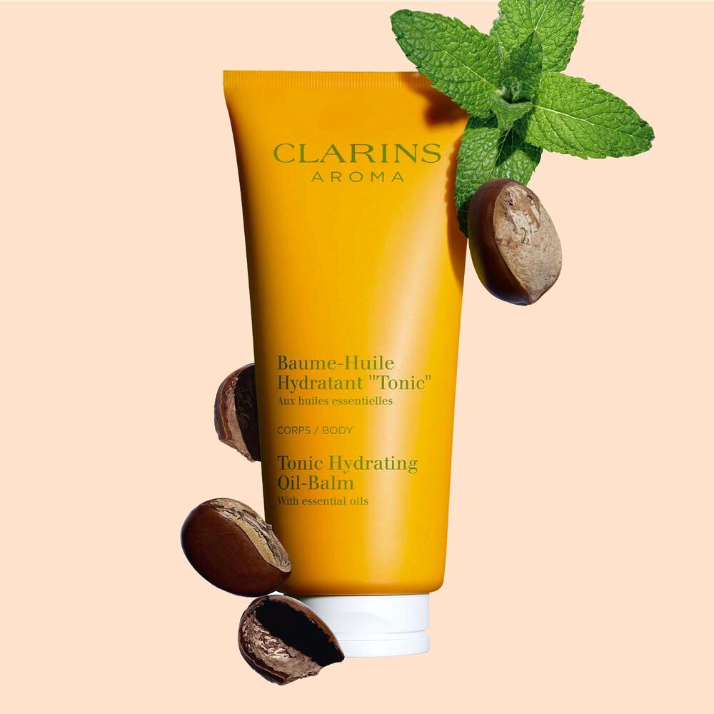 Plan produit d'un tube de Baume-Huile Hydratant "Tonic" Clarins, avec feuilles de menthe et graines de karité sur fond pêche.