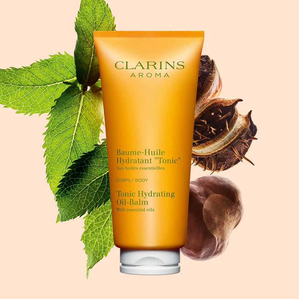 Packshot einer Tube Clarins Baume-Huile Tonic Öl-Balsam mit ein paar Minzeblättern und Sheanüssen vor einem pfirsichfarbenen Hintergrund.