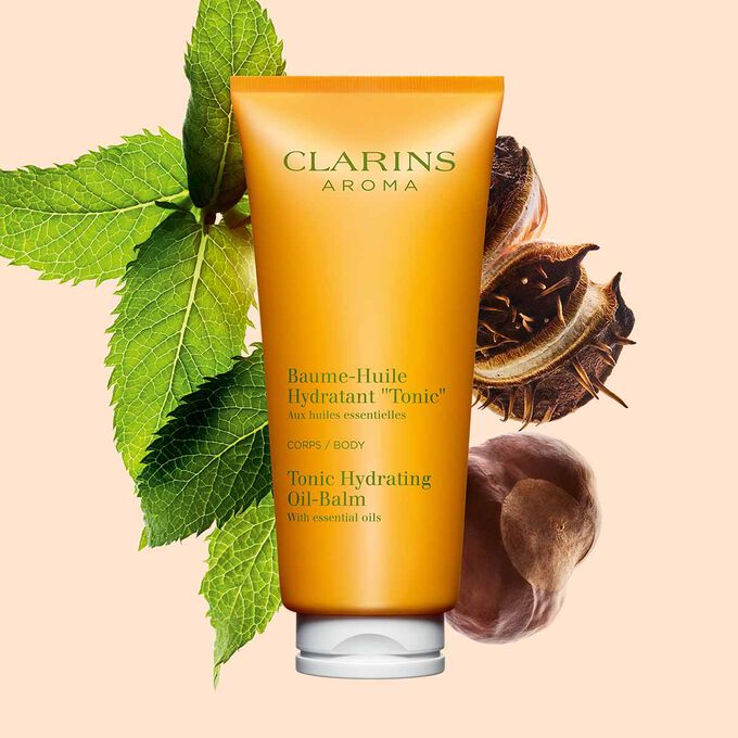 Packshot einer Tube Clarins Baume-Huile Tonic Öl-Balsam mit ein paar Minzeblättern und Sheanüssen vor einem pfirsichfarbenen Hintergrund.