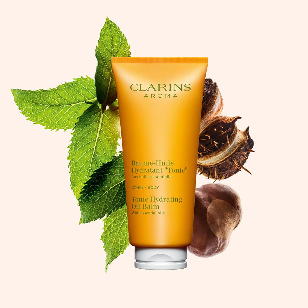 Packshot einer Tube Clarins Baume-Huile Tonic &Ouml;l-Balsam mit ein paar Minzebl&auml;ttern und Shean&uuml;ssen vor einem pfirsichfarbenen Hintergrund.