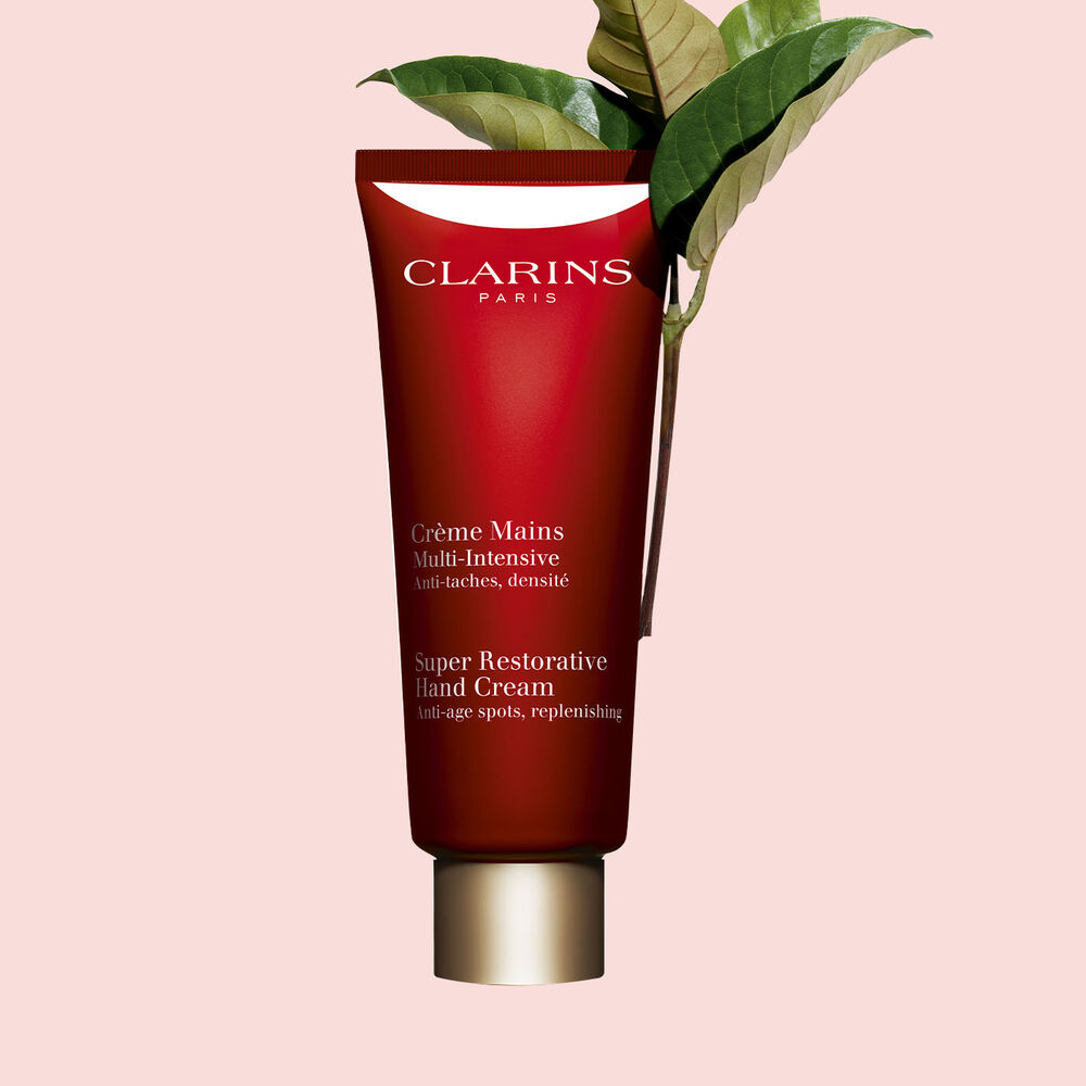 Packshot einer Tube Multi-Intensive Handcreme von Clarins mit ein paar Haronga-Blättern vor einem rosa Hintergrund.