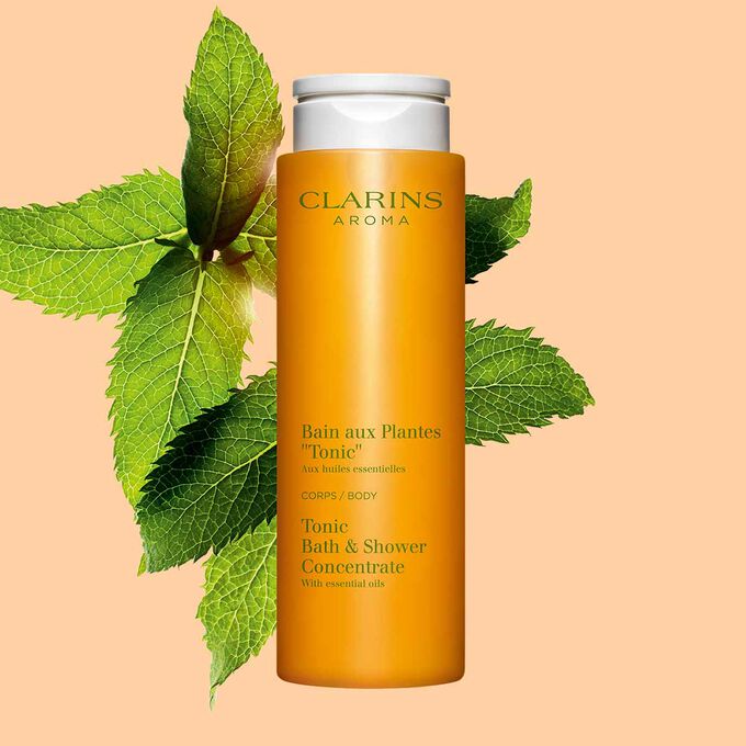 Packshot von Clarins Pflanzenbad Tonic mit einigen Minze- und Rosmarinblättern vor einem orangen Hintergrund.