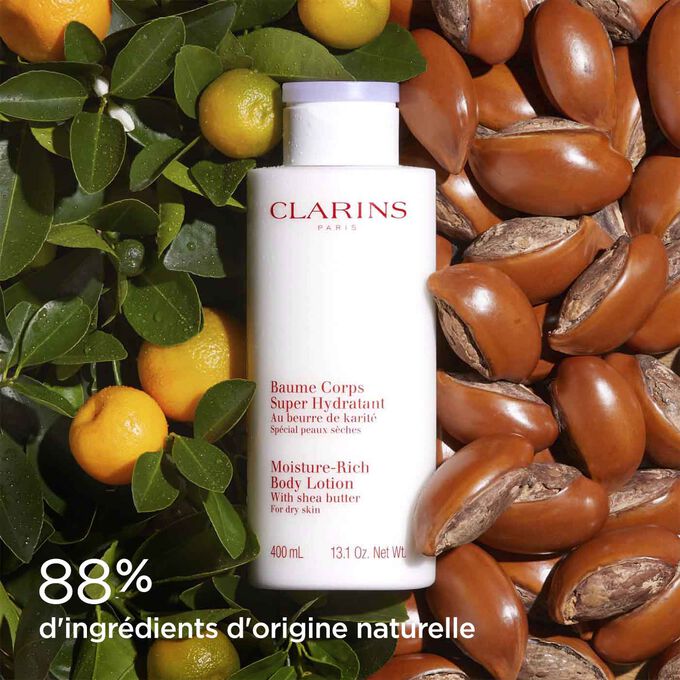 Flacon du Baume Corps Super Hydratant Clarins entouré de noix de karité et oranges, pour illustrer ses ingrédients naturels.